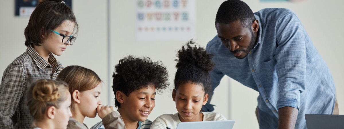 Diverse group of children with male teacher using laptop together in modern school classroom